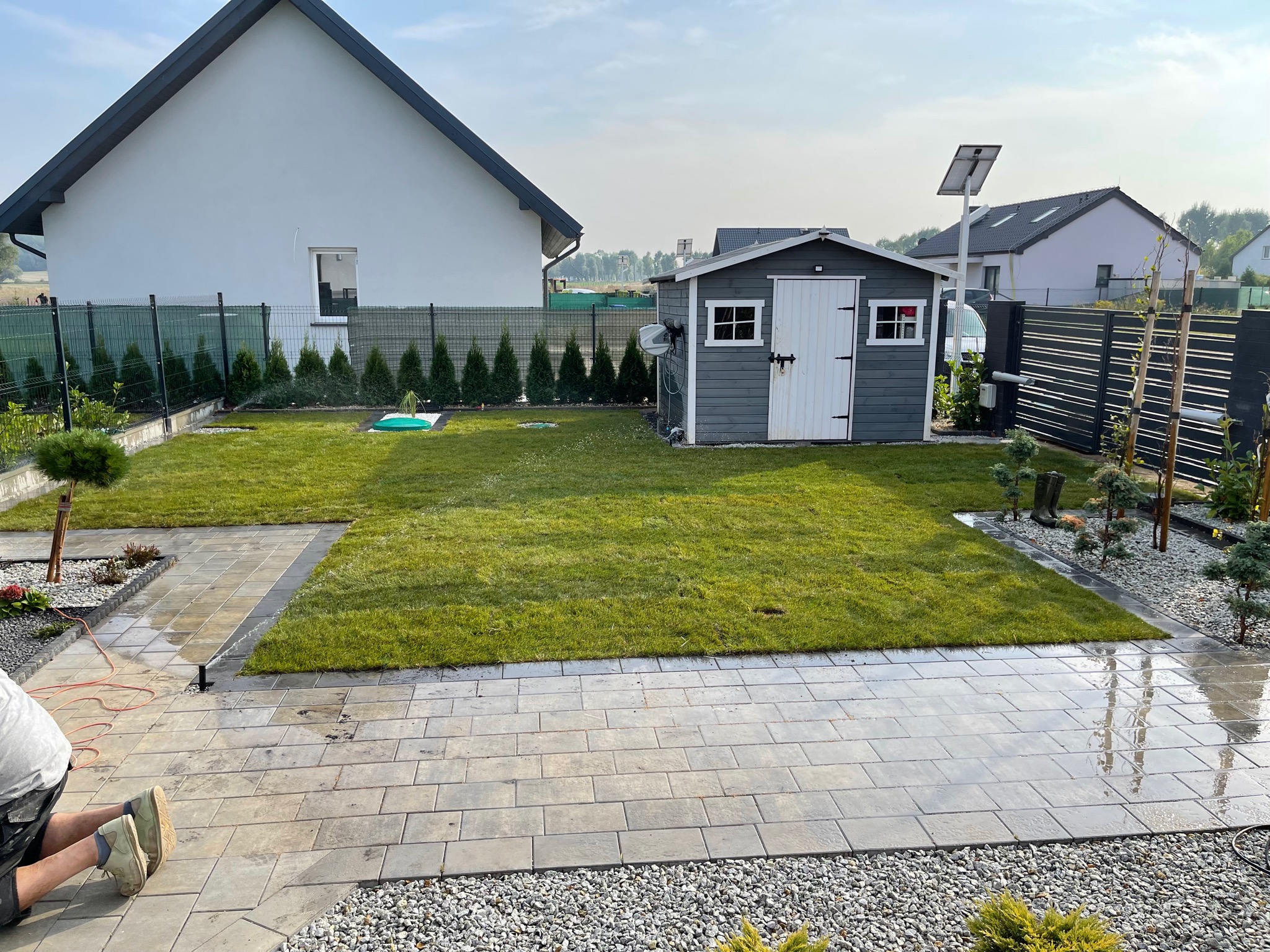 Świeżo założony trawnik z szarą altaną narzędziową i kostką brukową, widok z poziomu brukarza w roboczych butach, dom w tle.