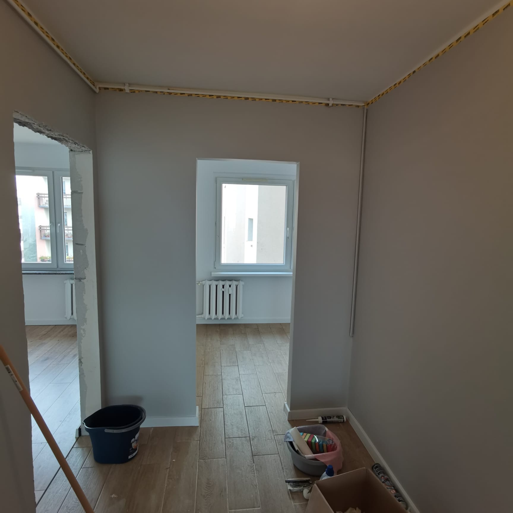 Przygotowanie do malowania ścian w szarym kolorze, widoczne zabezpieczenia taśmą malarską na suficie, wiadro i narzędzia malarskie na podłodze z płytek imitujących drewno.