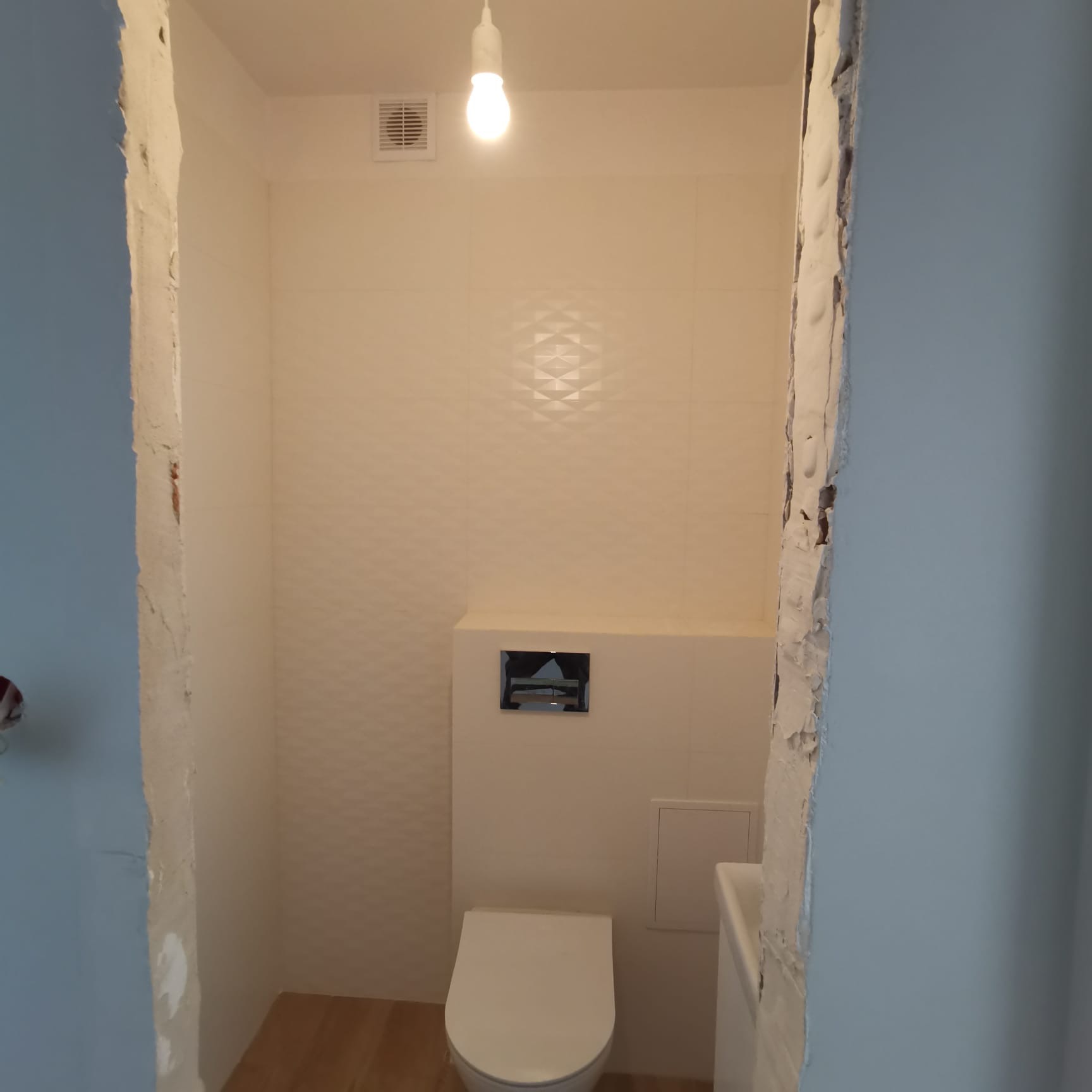 Wykończona toaleta z podwieszaną miską WC, z widocznymi fragmentami nieotynkowanych ścian po bokach, drewnopodobna podłoga i białe płytki z delikatnym wzorem na ścianach.