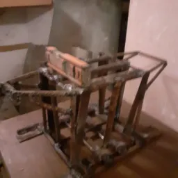 Metalowa konstrukcja klatki z widocznymi śladami spawania, leżąca na drewnianej powierzchni, z poziomicą na górze, sfotografowana w słabym oświetleniu.