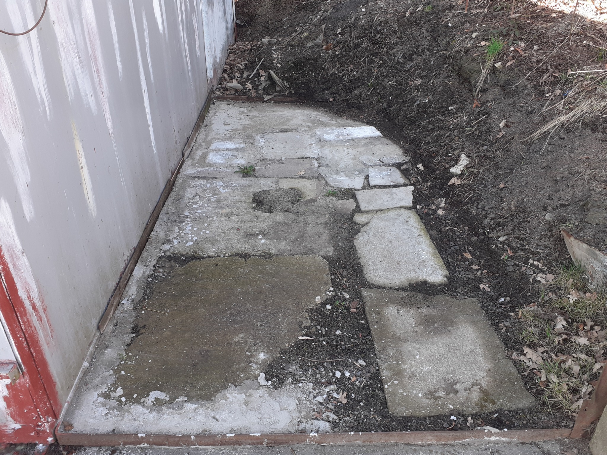 Nierówny betonowy chodnik z płyt przy ścianie z blachy falistej, widoczne ślady wilgoci i zabrudzenia, ziemia z liśćmi po prawej stronie.