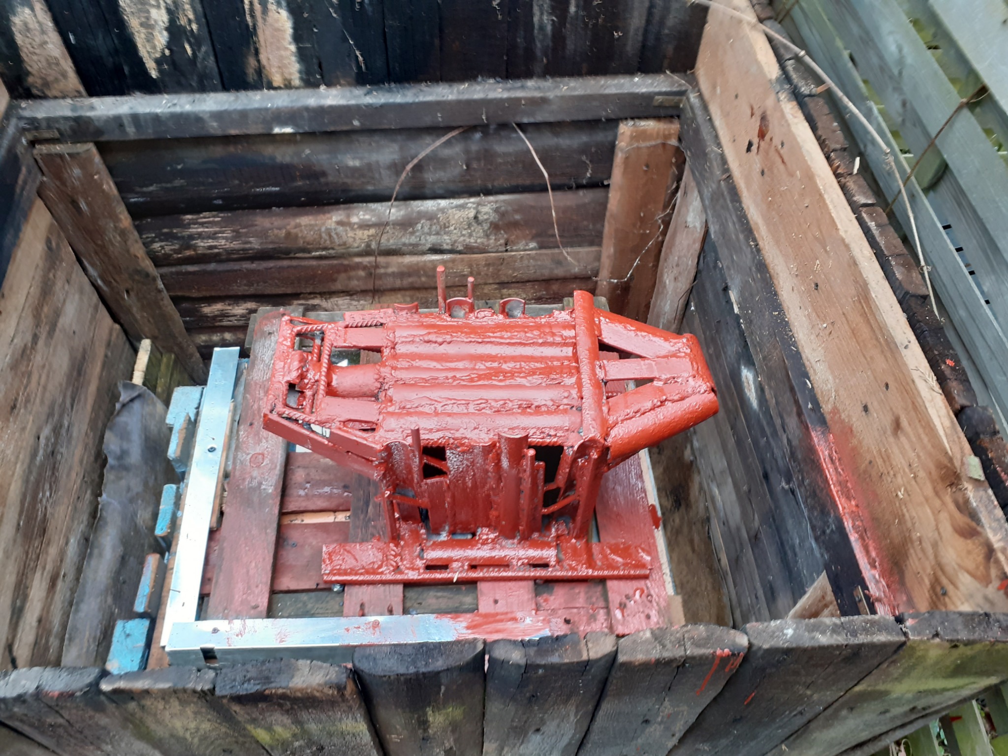 Czerwona konstrukcja stalowa, świeżo pomalowana, umieszczona w drewnianej skrzyni, widoczne ślady farby na drewnie.