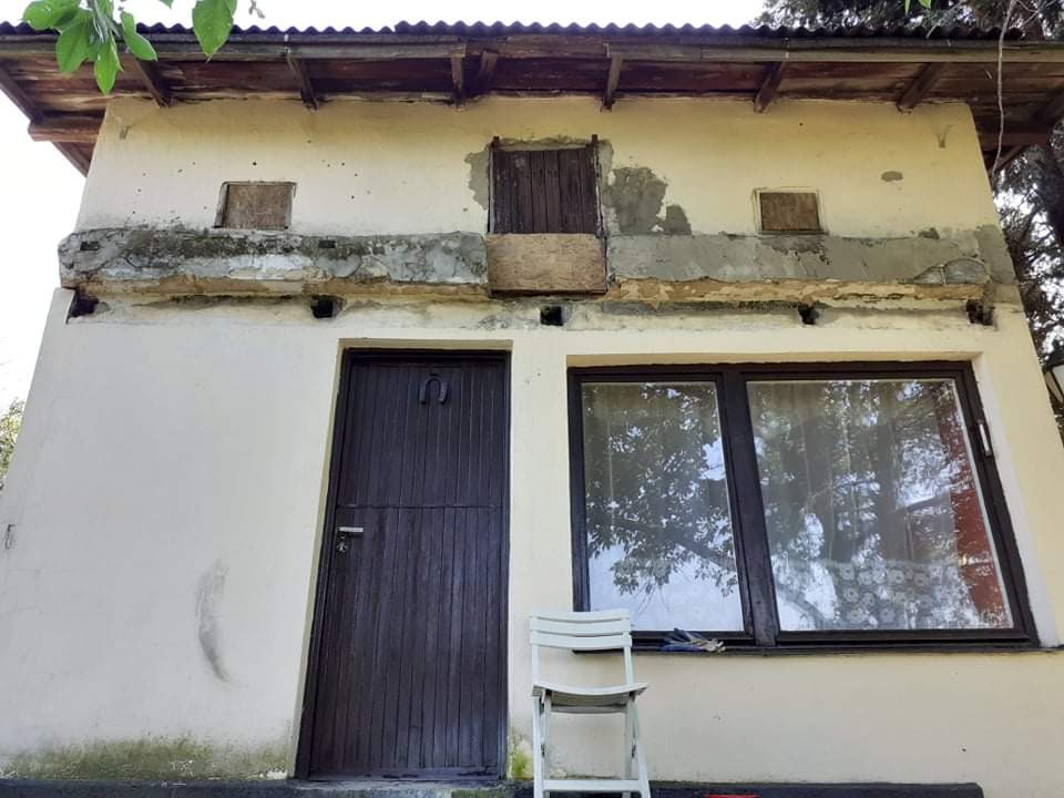 Dwukondygnacyjny budynek z widocznymi śladami napraw i remontów, z oknem i drzwiami w ciemnej ramie, białe krzesło przed oknem, zabetonowany fragment elewacji na górze.