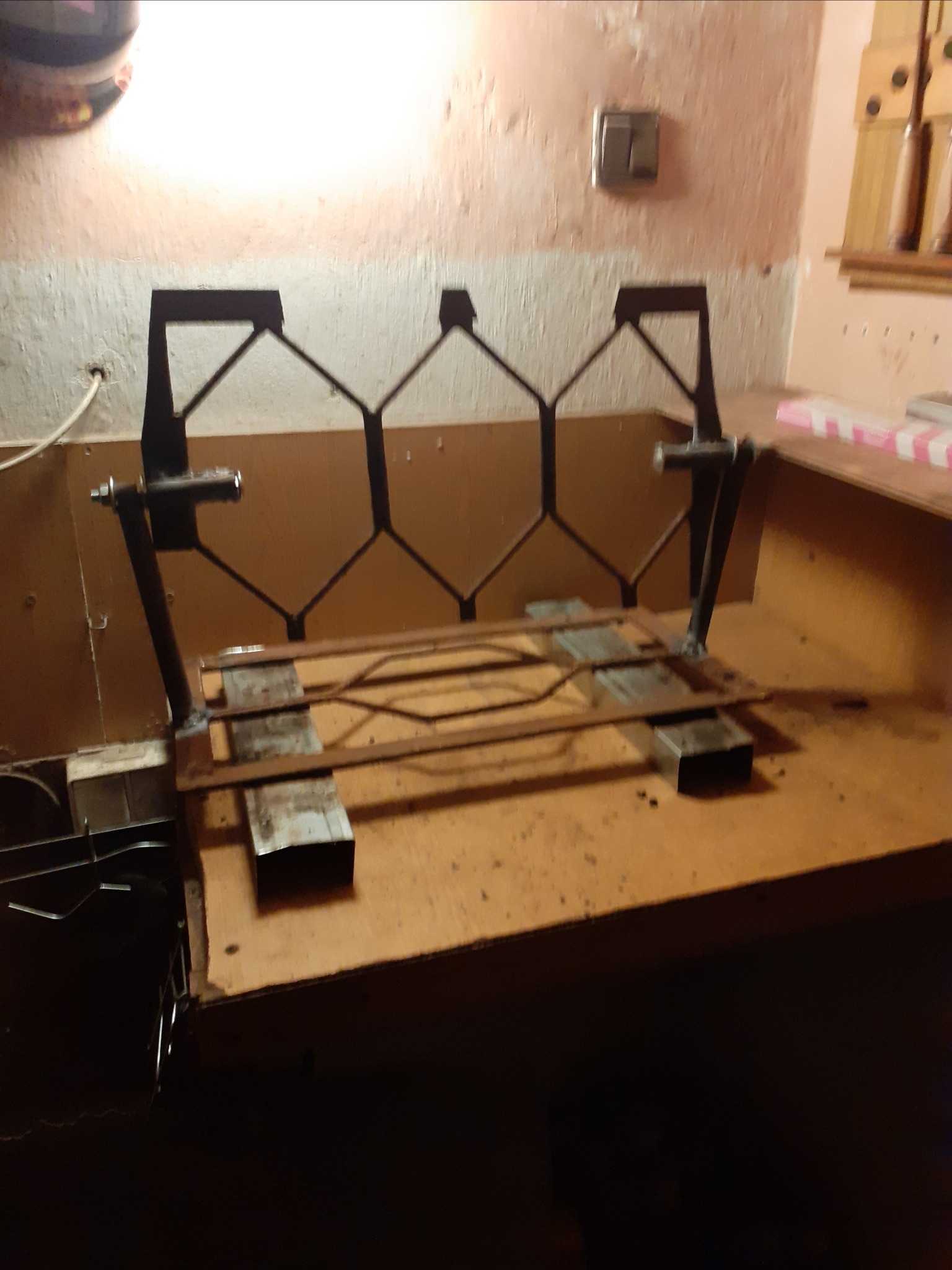 Surowa, niedokończona konstrukcja z metalowych profili, z widocznymi spawami i śladami rdzy, umieszczona na warsztatowym stole w słabo oświetlonym pomieszczeniu.