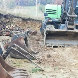 Zielona koparka kołowa z kilkoma różnymi łyżkami, leżącymi na ziemi, na tle wykopu i sterty pociętego drewna.