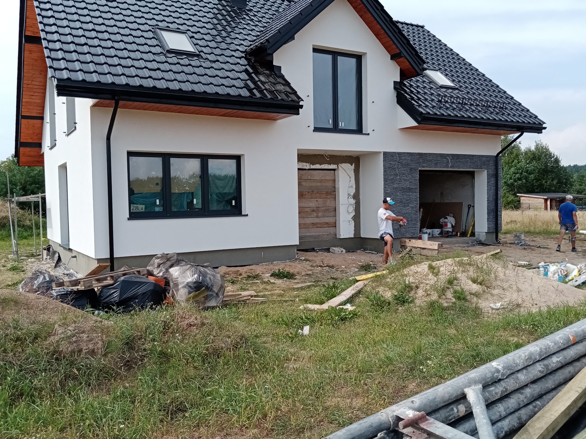 Dwukondygnacyjny dom w trakcie prac elewacyjnych, z widocznym ociepleniem i szpachlowaniem, fragment elewacji wykończony szarym kamieniem, na pierwszym planie materiały budowlane i fragment...