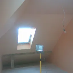 Poddasze w Częstochowie po adaptacji, widoczne okno dachowe, ściany przygotowane pod malowanie, surowa żarówka zwisająca z sufitu oraz lampa robocza na statywie.