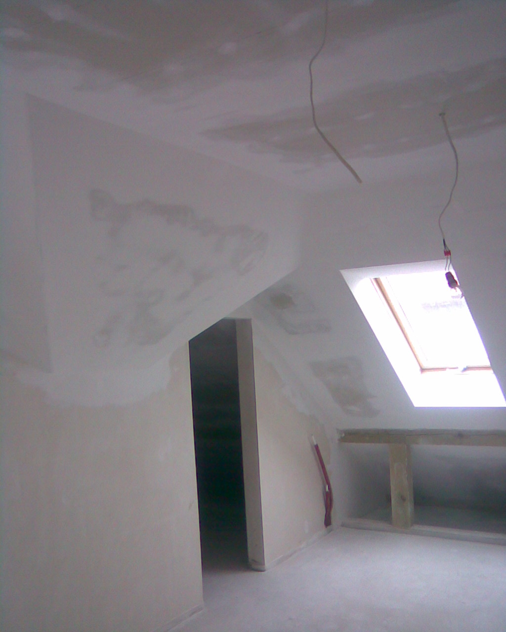 Poddasze w trakcie adaptacji: ściany i sufit po wstępnym szpachlowaniu, widoczne ślady po łatach, okno dachowe w skosie, wystające przewody elektryczne.