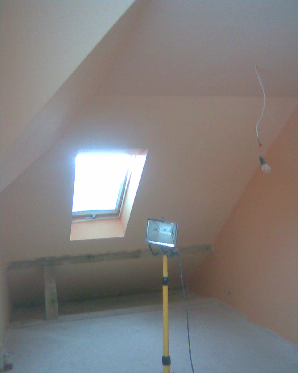 Poddasze w Częstochowie po adaptacji, widoczne okno dachowe, ściany przygotowane pod malowanie, surowa żarówka zwisająca z sufitu oraz lampa robocza na statywie.