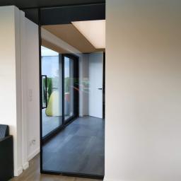 AD-BUD LENA DUB - Nowoczesne wnętrze z przesuwnymi drzwiami szklanymi w czarnej ramie, szare płytki podłogowe, białe ściany i ciemny sufit, widok na ogród przez okno.