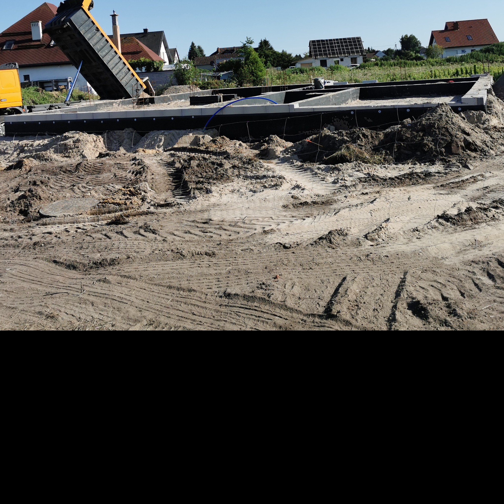 Wyładunek piasku z wywrotki na przygotowany fundament domu jednorodzinnego z widoczną hydroizolacją i zbrojeniami.