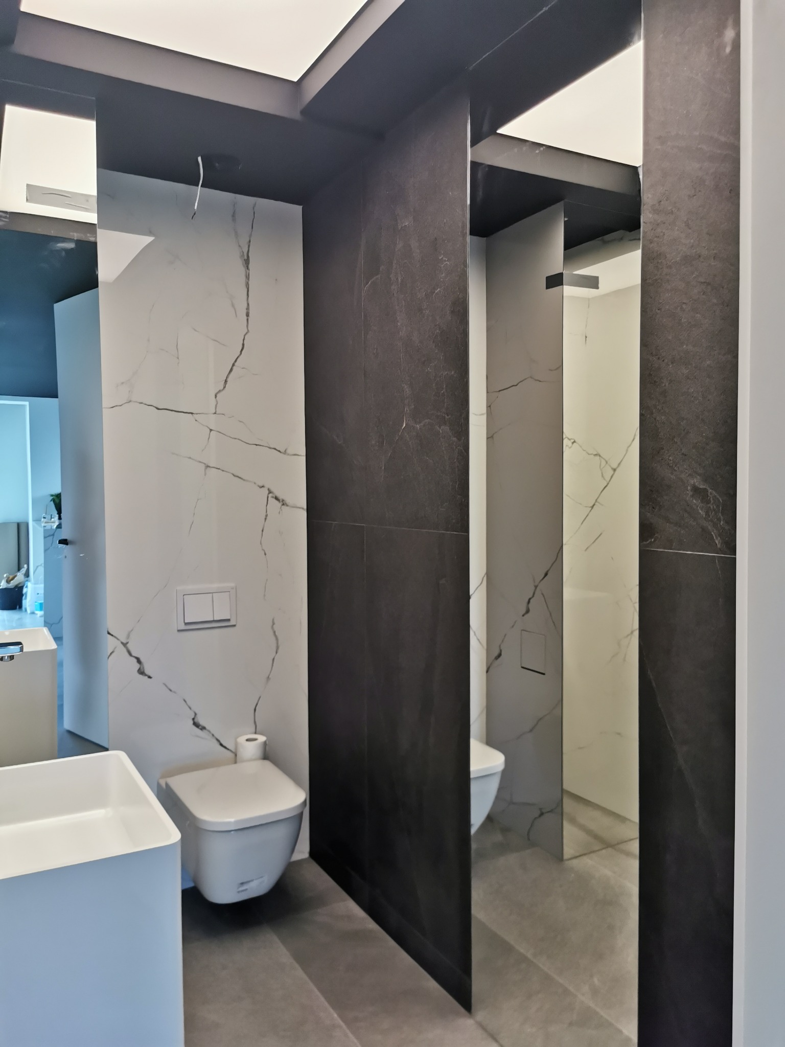 Nowoczesna łazienka z białymi płytkami imitującymi marmur i ciemnoszarymi płytkami, wisząca miska WC z podwójnym przyciskiem spłukującym, biała umywalka nablatowa, szara podłoga i oświetlenie...