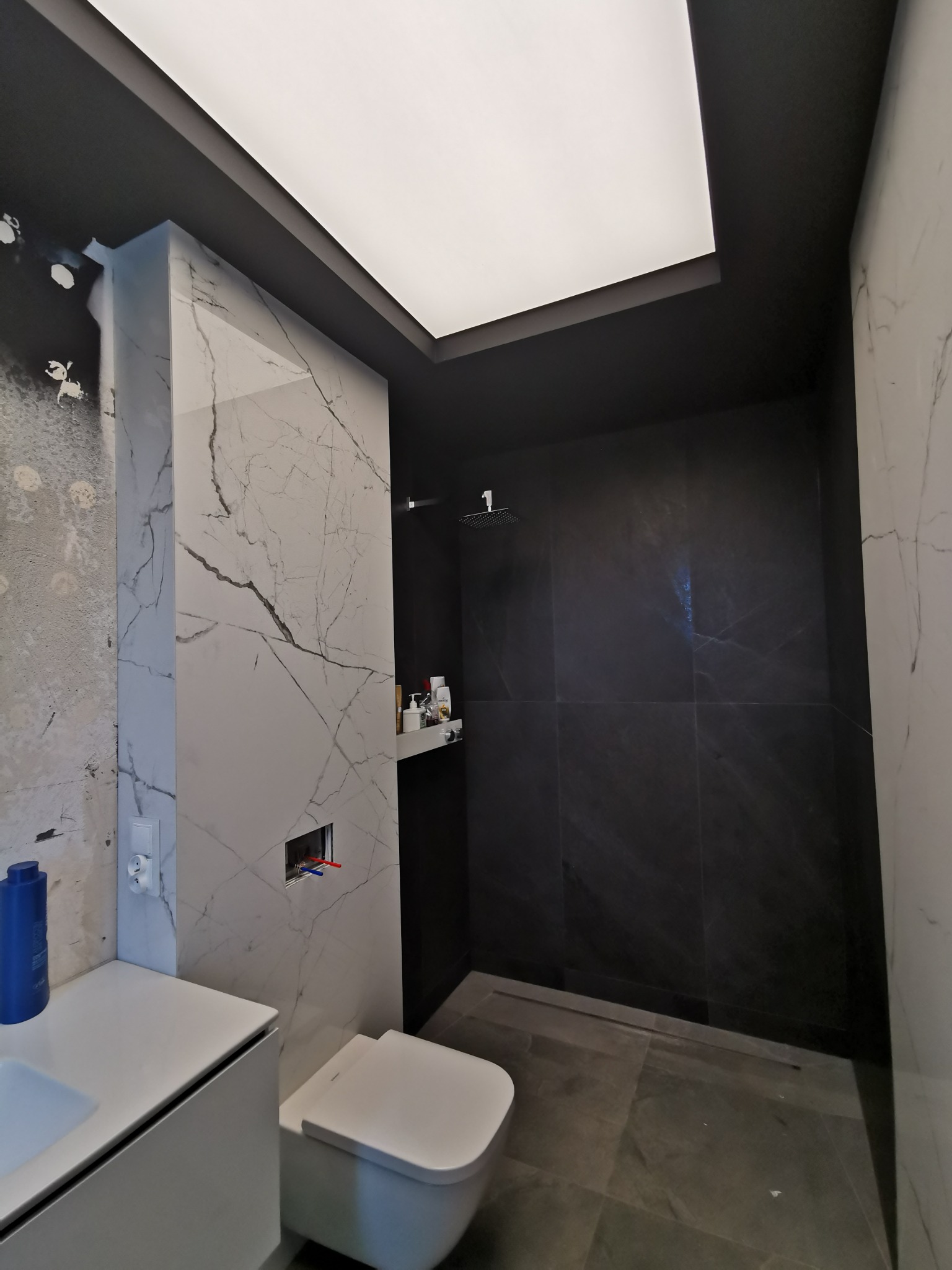 Wnętrze łazienki z czarną kabiną prysznicową i białymi płytkami imitującymi marmur, widoczna wisząca toaleta i umywalka z szafką, sufit podwieszany z oświetleniem LED.