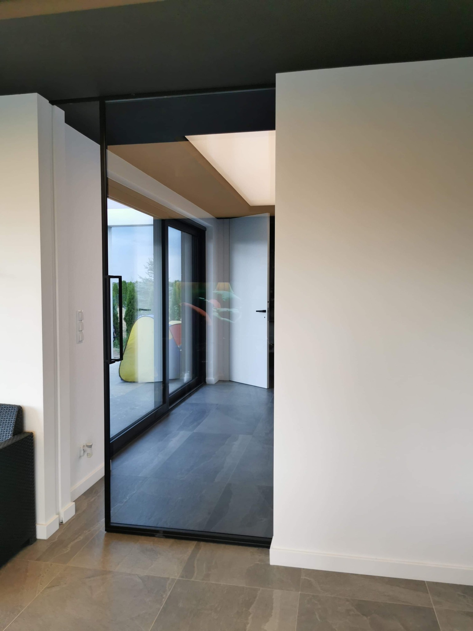 Nowoczesne wnętrze z przesuwnymi drzwiami szklanymi w czarnej ramie, szare płytki podłogowe, białe ściany i ciemny sufit, widok na ogród przez okno.