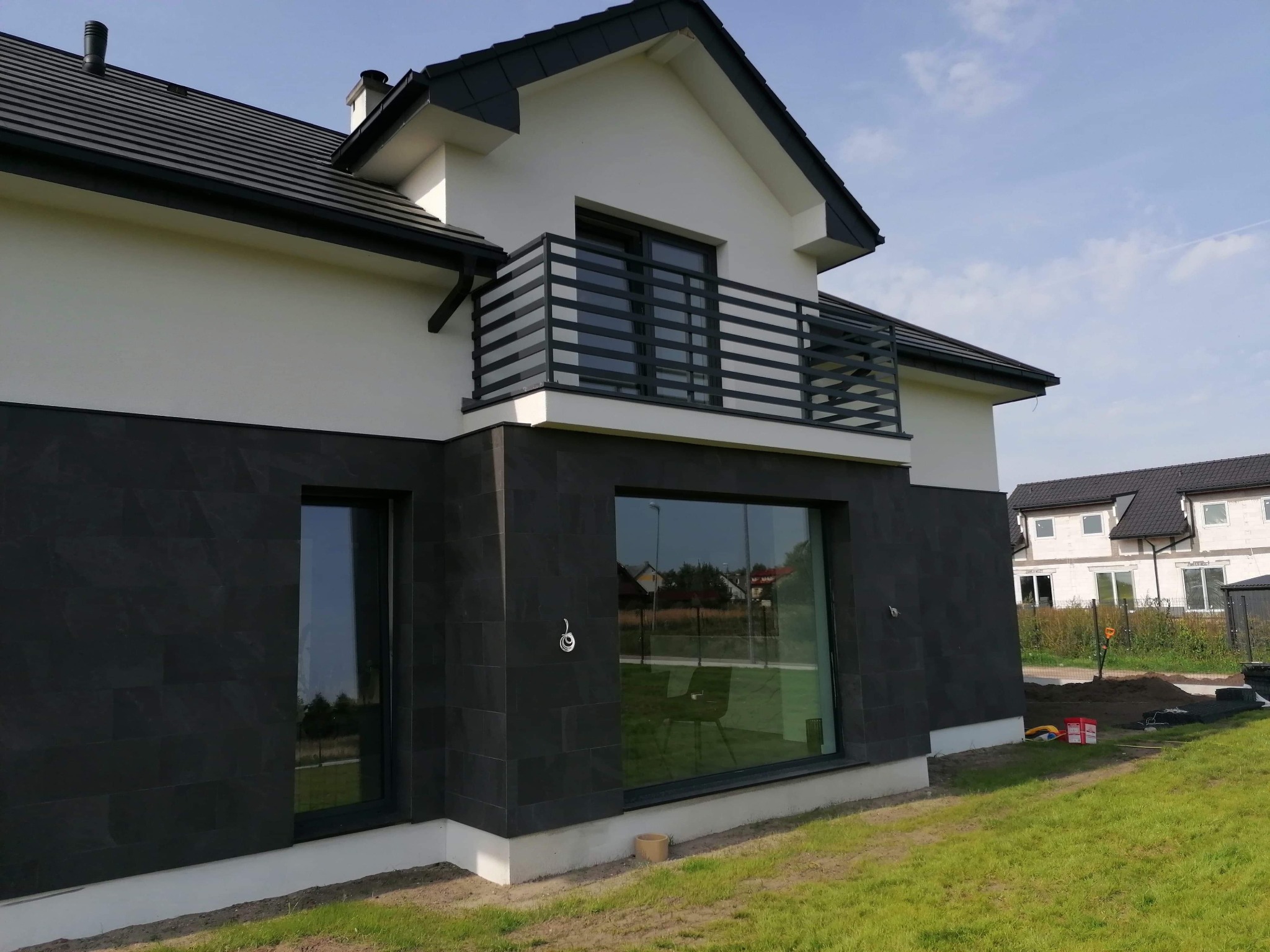 Nowoczesny dom jednorodzinny z balkonem i elewacją wykończoną ciemnymi płytkami imitującymi kamień, widoczne okna z czarnymi ramami i świeżo założony trawnik.