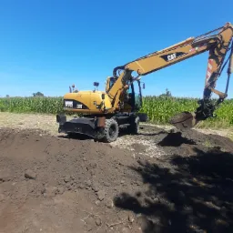 Żółta koparka CAT M315C na tle pola kukurydzy, wykonująca wykop ziemny w słoneczny dzień.