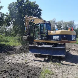 Żółto-czarna koparka CAT M315C nabiera ziemię na placu budowy w słoneczny dzień, widoczne drzewa w tle.