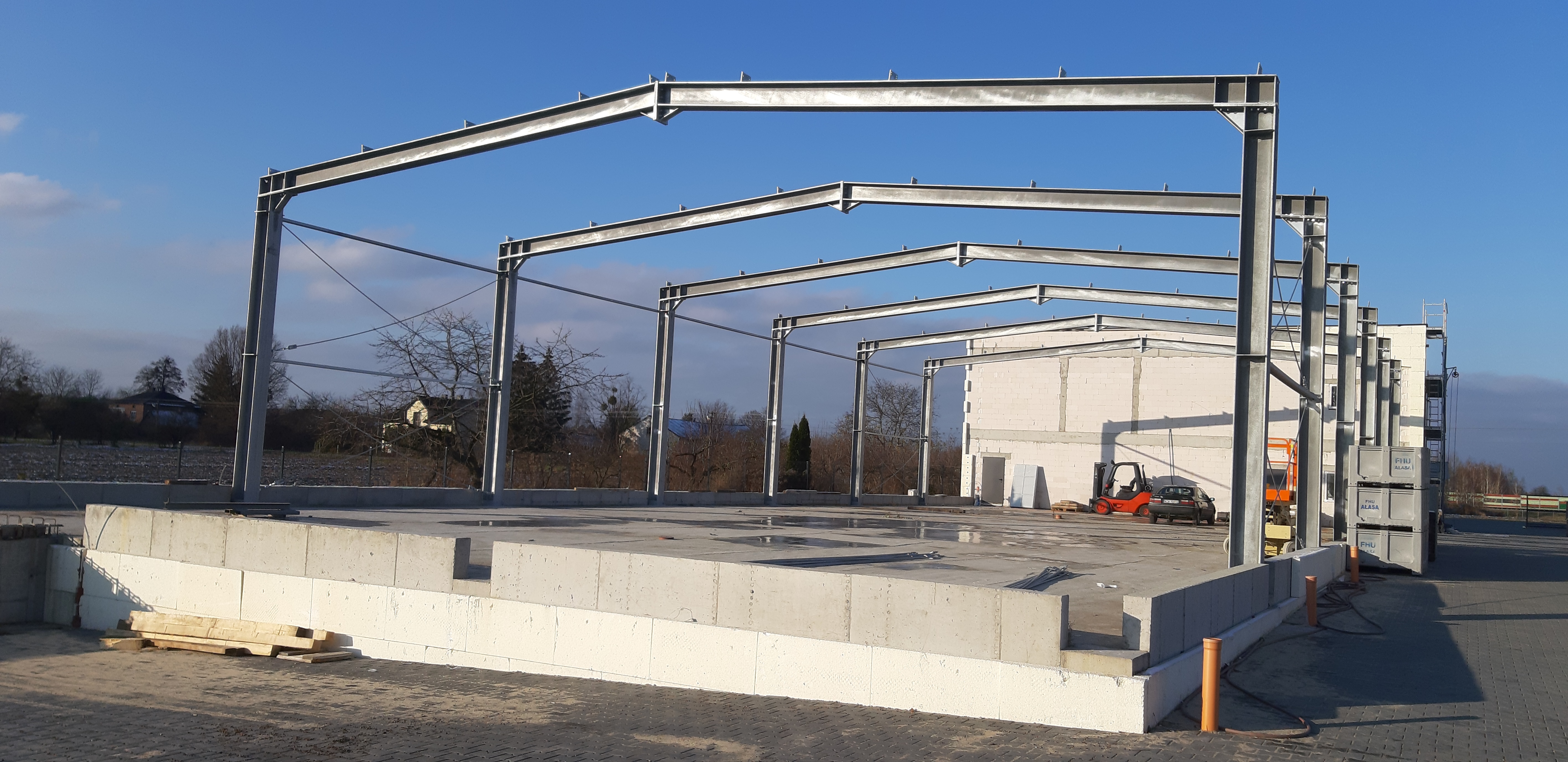 Montaż szkieletu stalowego hali na betonowej płycie fundamentowej, widoczny wózek widłowy i budynek w tle, słoneczny dzień.