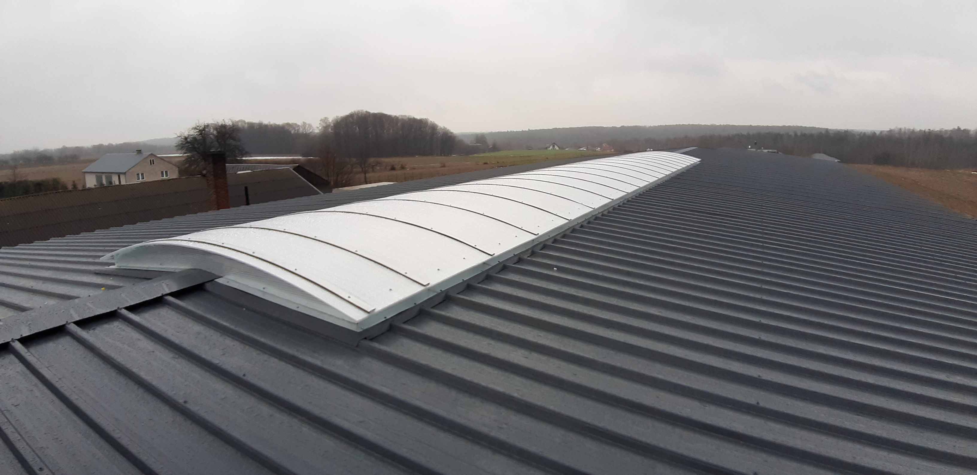 Szary dach z blachy trapezowej z zamontowanym świetlikiem łukowym, widok z góry na tle pochmurnego nieba i zabudowań.