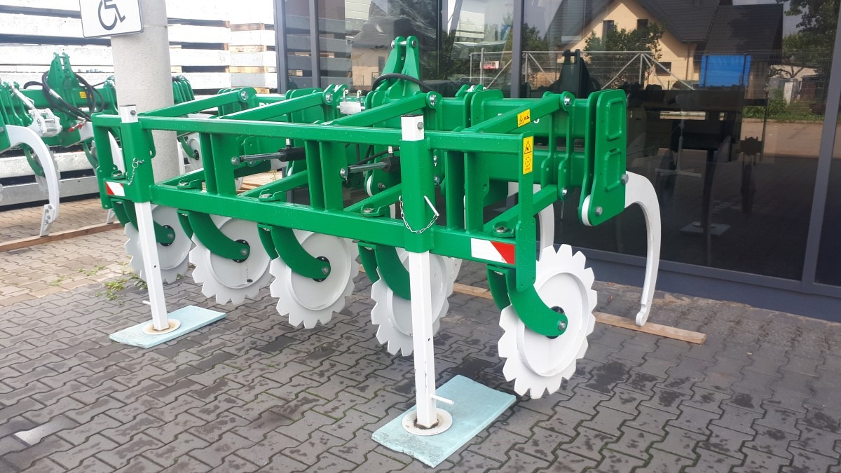 Zielona rama z białymi zębatymi tarczami, prawdopodobnie element maszyny rolniczej, stojąca na betonowej kostce brukowej przed budynkiem.