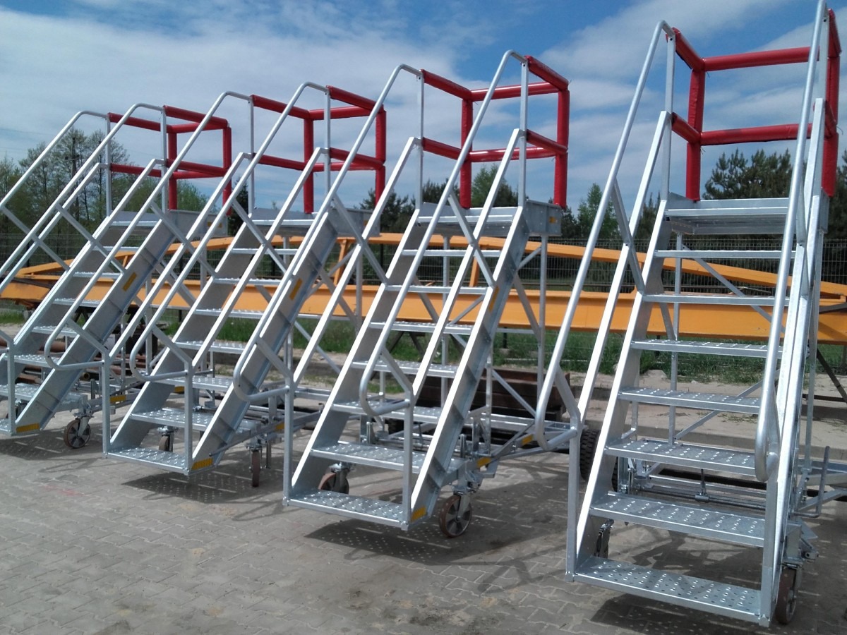 Mobilne schody metalowe z platformą, ocynkowane, z czerwoną barierką ochronną, na tle błękitnego nieba i zieleni drzew.