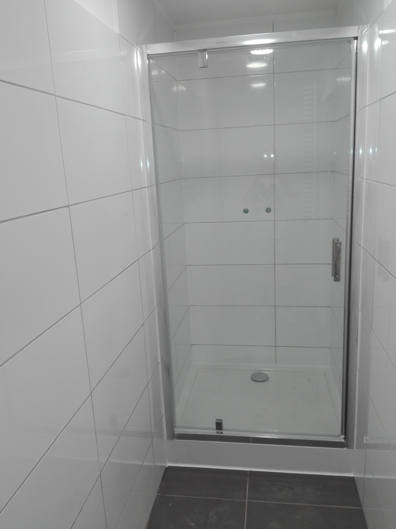 Nowa kabina prysznicowa z przesuwnymi drzwiami w łazience wyłożonej białymi, błyszczącymi płytkami kwadratowymi i ciemnoszarą podłogą.