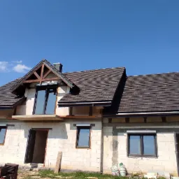 Dom w trakcie budowy z brązowym dachem z dachówki, widoczne okna z szarymi ramami, ściany z białego bloczka, na pierwszym planie paleta z dachówką.