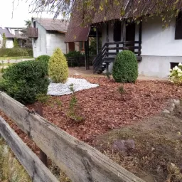 Ogród po renowacji: świeżo posadzone krzewy i drzewka ozdobne, kora rozsypana na rabatach, drewniany płot w rustykalnym stylu.