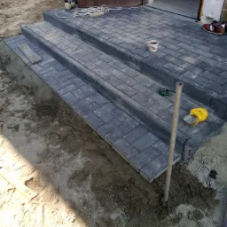 Budowa schodów wejściowych z szarej kostki brukowej, widoczne narzędzia pracy, poziomica, sznurek i kubek z kawą na gotowych stopniach.