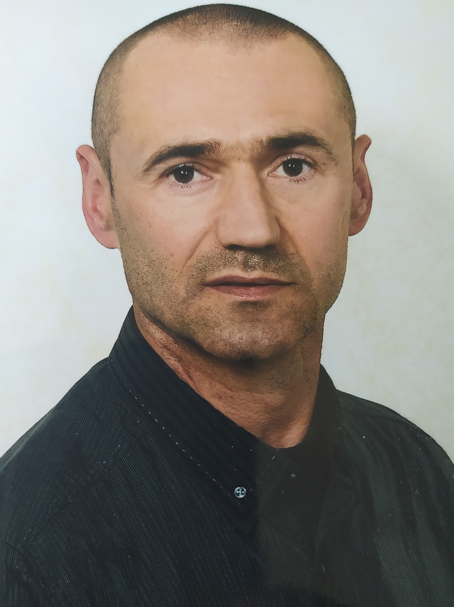 Portret mężczyzny w ciemnej koszuli z guzikami, krótko ostrzyżonego, z lekkim zarostem, na jasnym tle.