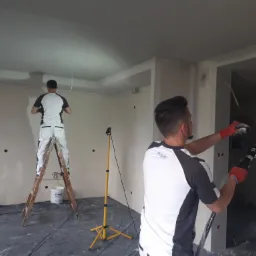 Dwóch mężczyzn w trakcie natryskowego nakładania gładzi na ściany w nowo wykończonym pomieszczeniu. Jeden stoi na drewnianej drabinie, drugi obsługuje maszynę natryskową w czerwonych rękawicach.