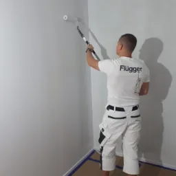 Mężczyzna w białej koszulce z logo Flűgger maluje ścianę na biało wałkiem, zabezpieczone niebieską taśmą narożniki, widok z tyłu.