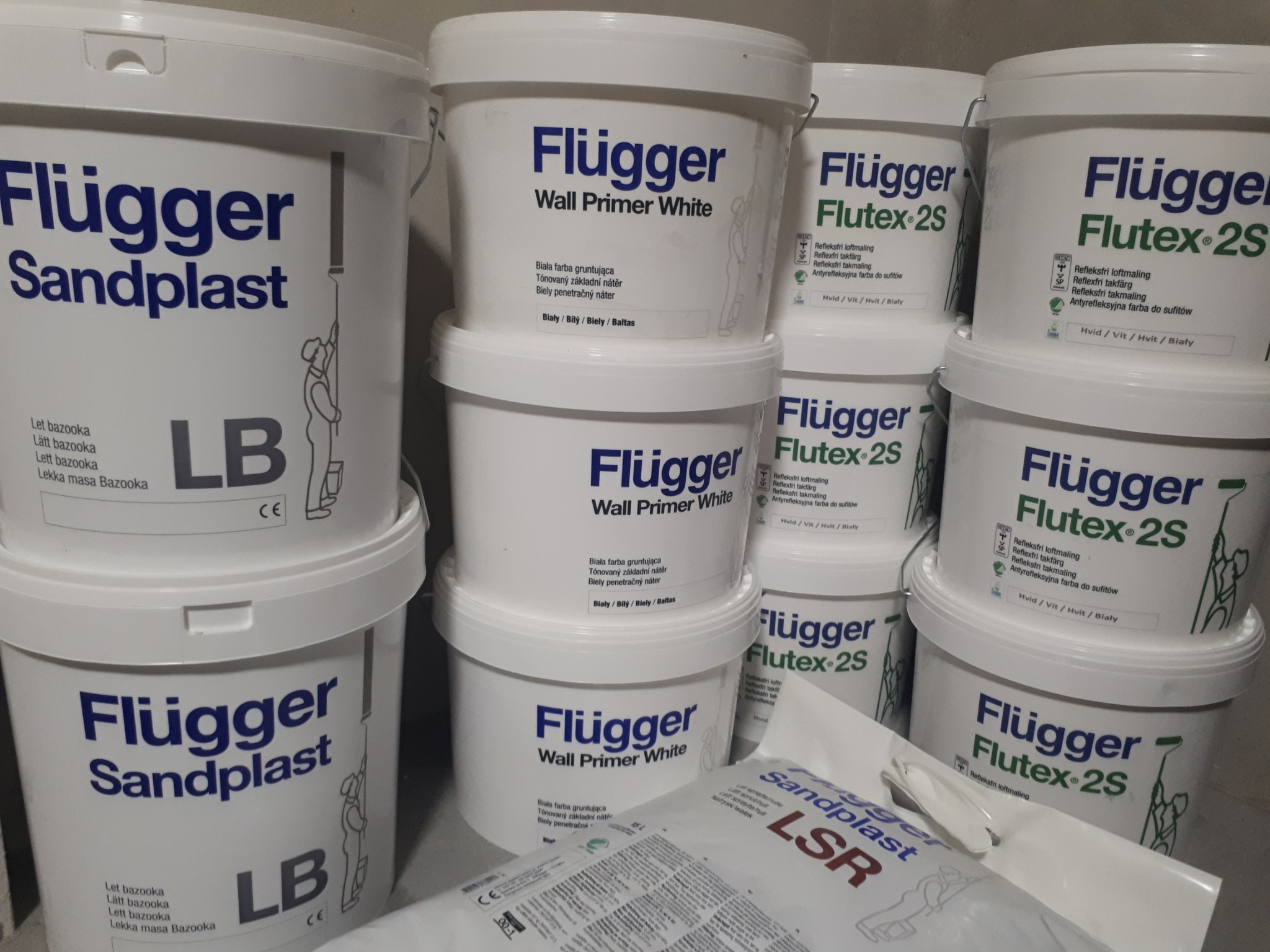 Stos białych wiader z farbami i gruntami marki Flugger: Sandplast, Wall Primer White i Flutex 2S, widoczne również opakowania uzupełniające Sandplast LSR.