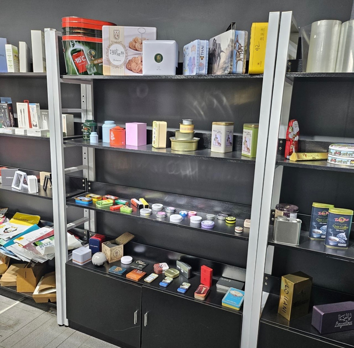 Półki ekspozycyjne w sklepie lub magazynie z różnorodnymi metalowymi i kartonowymi pudełkami i puszkami, niektóre z nadrukami w różnych kolorach i wzorach, w tym puszki na kawę i słodycze.