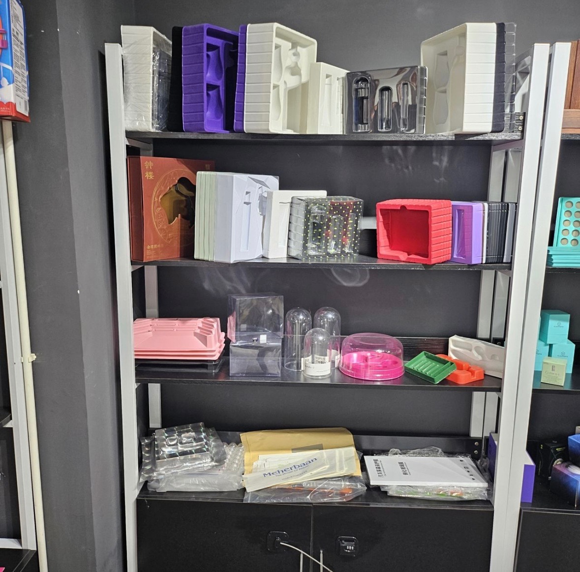 Regał z różnorodnymi opakowaniami z tworzyw sztucznych, w tym pudełka z przegródkami, przezroczyste kopuły i tacki, z widocznym logo 'Meherbaan' na jednym z opakowań na dolnej półce.