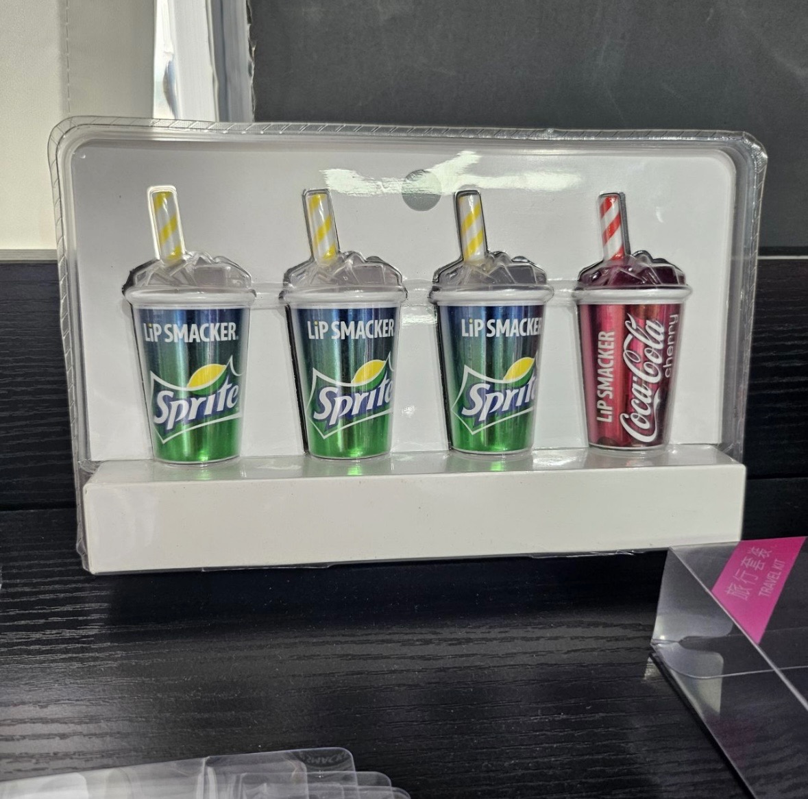 Zestaw czterech balsamów do ust Lip Smacker w kształcie kubków napojów Sprite i Coca-Cola Cherry, zapakowanych w przezroczyste pudełko na białym podłożu.