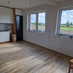 BEST-BUD - Jasne drewnopodobne panele podłogowe w nowym wnętrzu, widok na okna z widokiem na krajobraz, fragment zabudowy kuchennej z lodówką i kartonem po sprzęcie AGD.