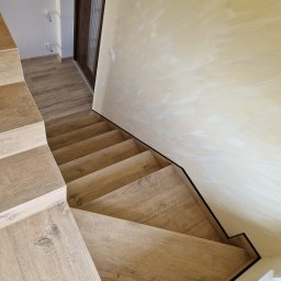 BEST-BUD - Drewniane schody z panelami podłogowymi na stopniach, kremowa ściana z delikatną strukturą, widok z góry, listwa przypodłogowa w kolorze czarnym.