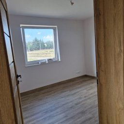 BEST-BUD - Pusty, jasny pokój z drewnianą podłogą i świeżo pomalowanymi ścianami, widok przez otwarte, drewniane drzwi, okno z widokiem na zewnątrz, przygotowany do dalszych prac wykończeniowych.