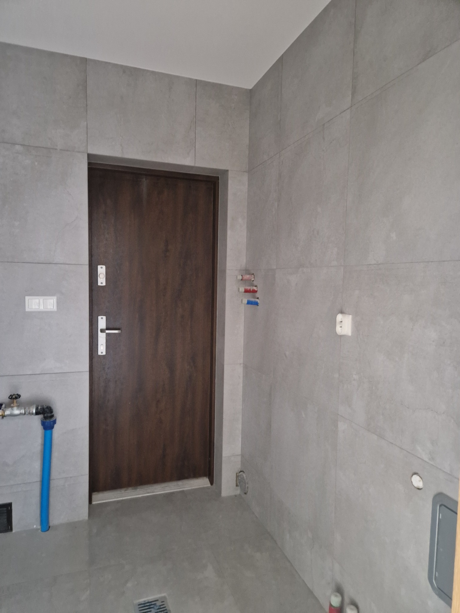 Wykończona łazienka z szarymi płytkami wielkoformatowymi na ścianach i podłodze, brązowe drzwi wejściowe z metalową klamką, widoczne elementy instalacji hydraulicznej i elektrycznej, kratka...