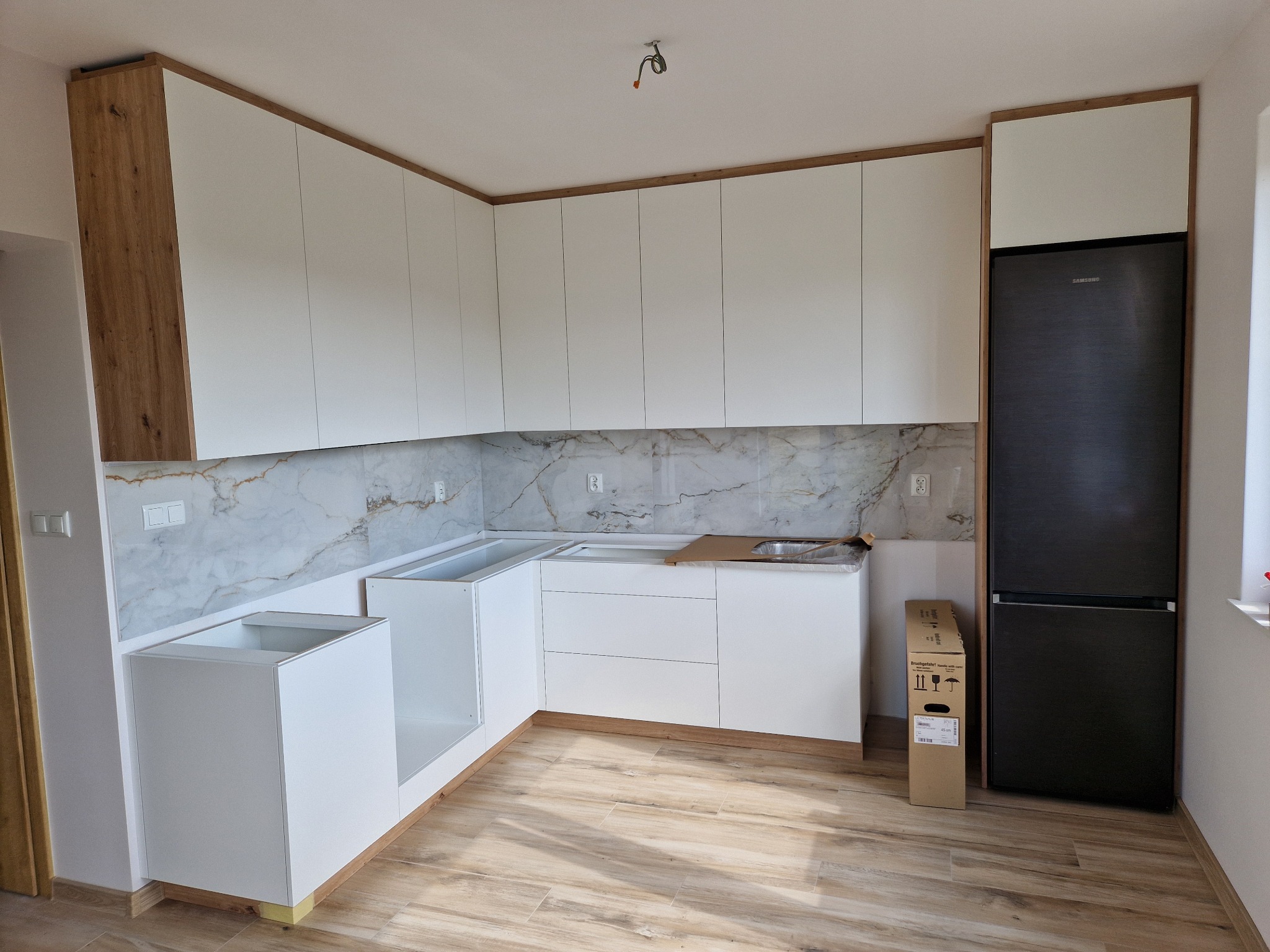 Nowoczesna, jasna kuchnia w trakcie montażu w Łomży: białe szafki z drewnianymi akcentami, marmurowy backsplash, karton po sprzęcie AGD obok lodówki.