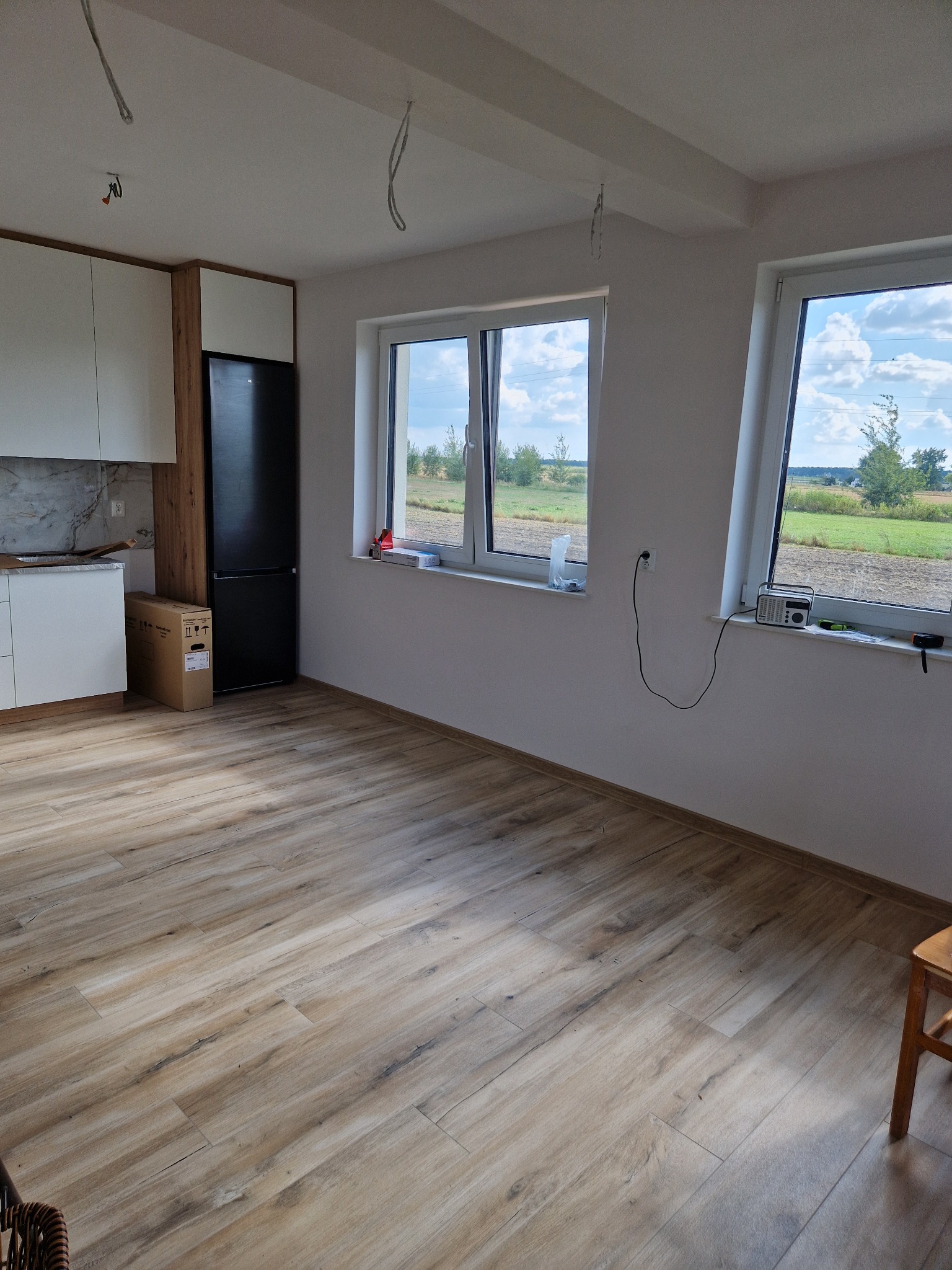 Jasne drewnopodobne panele podłogowe w nowym wnętrzu, widok na okna z widokiem na krajobraz, fragment zabudowy kuchennej z lodówką i kartonem po sprzęcie AGD.