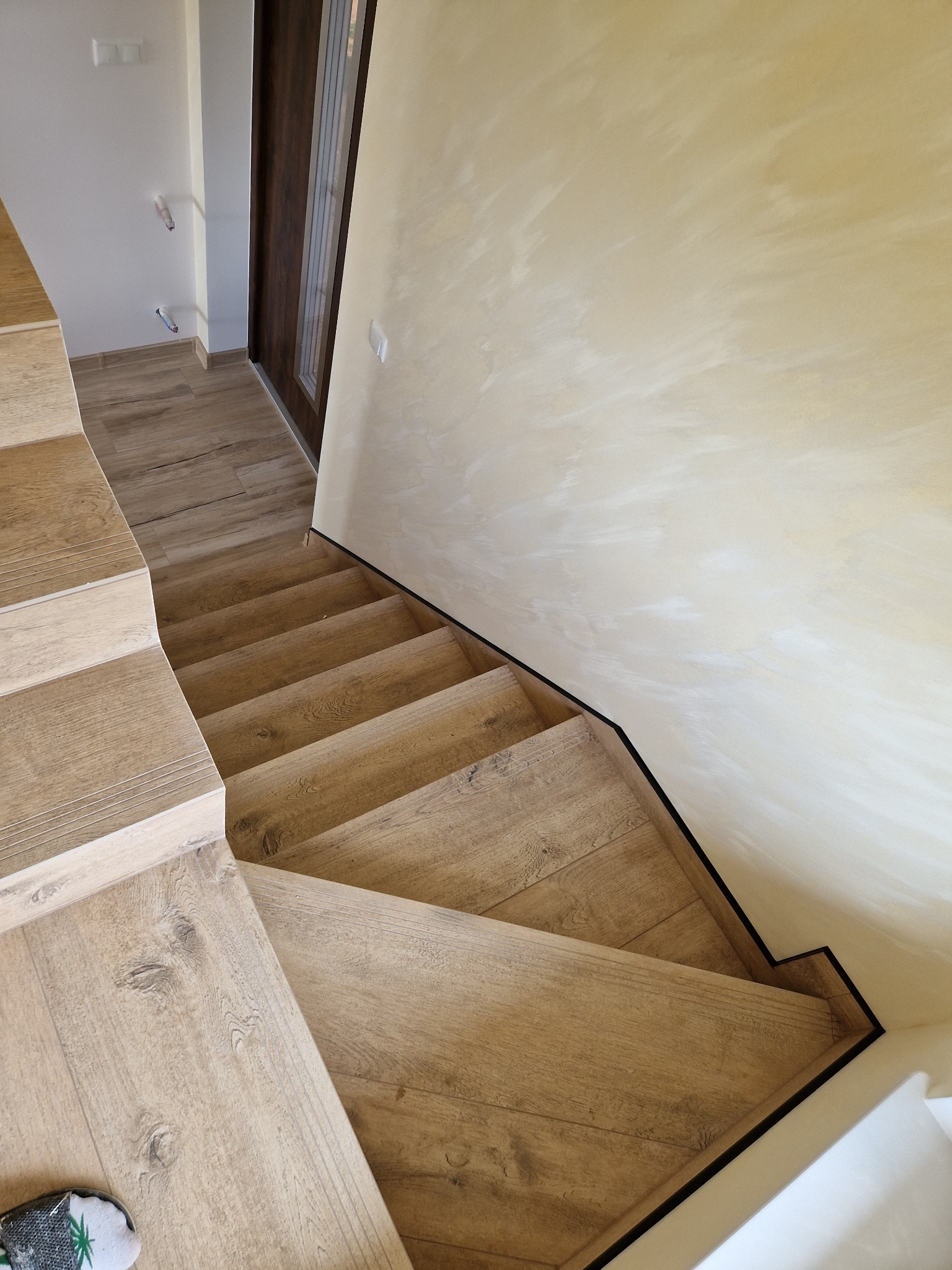 Drewniane schody z panelami podłogowymi na stopniach, kremowa ściana z delikatną strukturą, widok z góry, listwa przypodłogowa w kolorze czarnym.