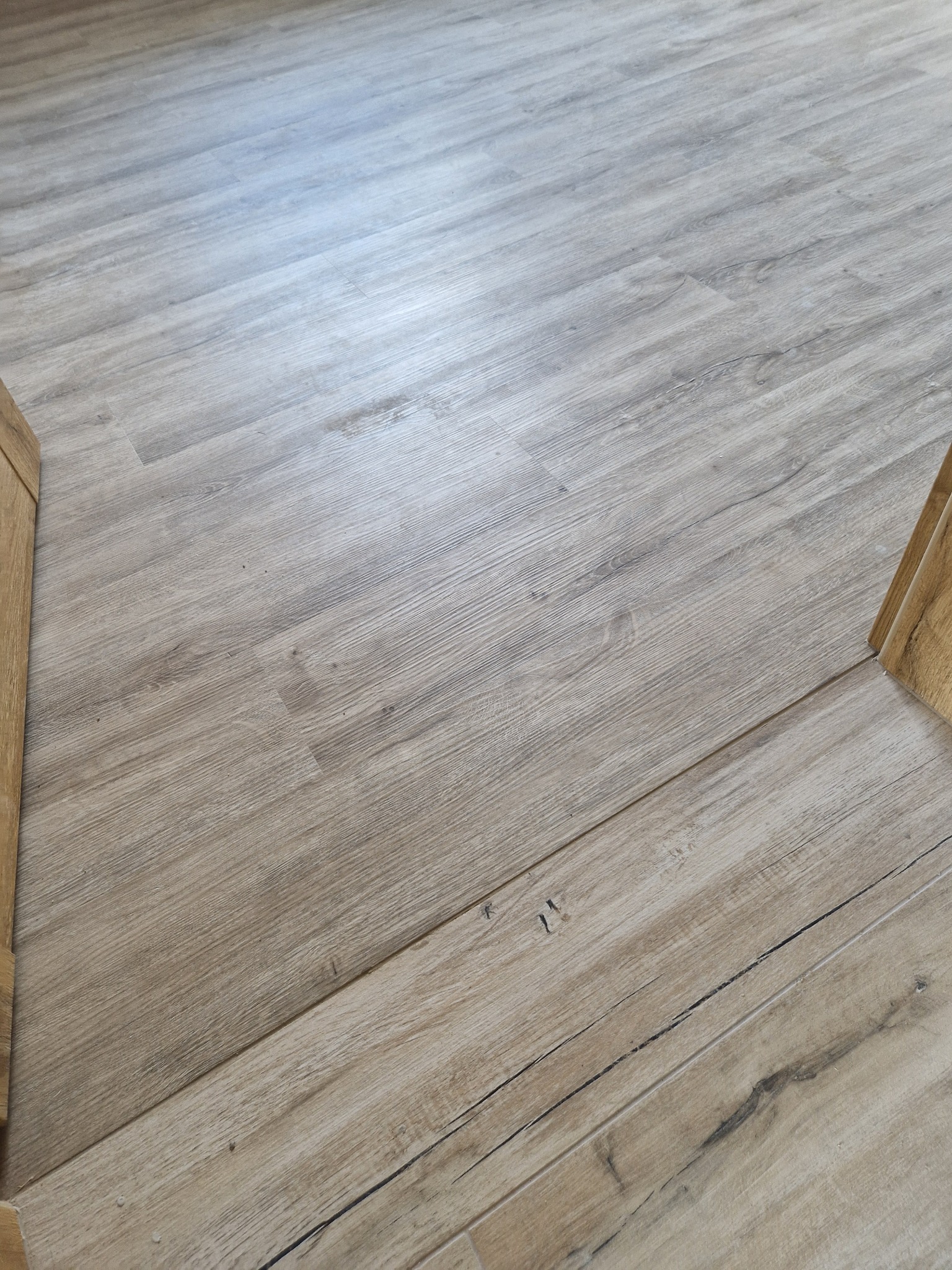 Jasna, drewnopodobna podłoga z widoczną strukturą słojów, ułożona pod kątem, z fragmentami przypodłogowych listew po bokach.