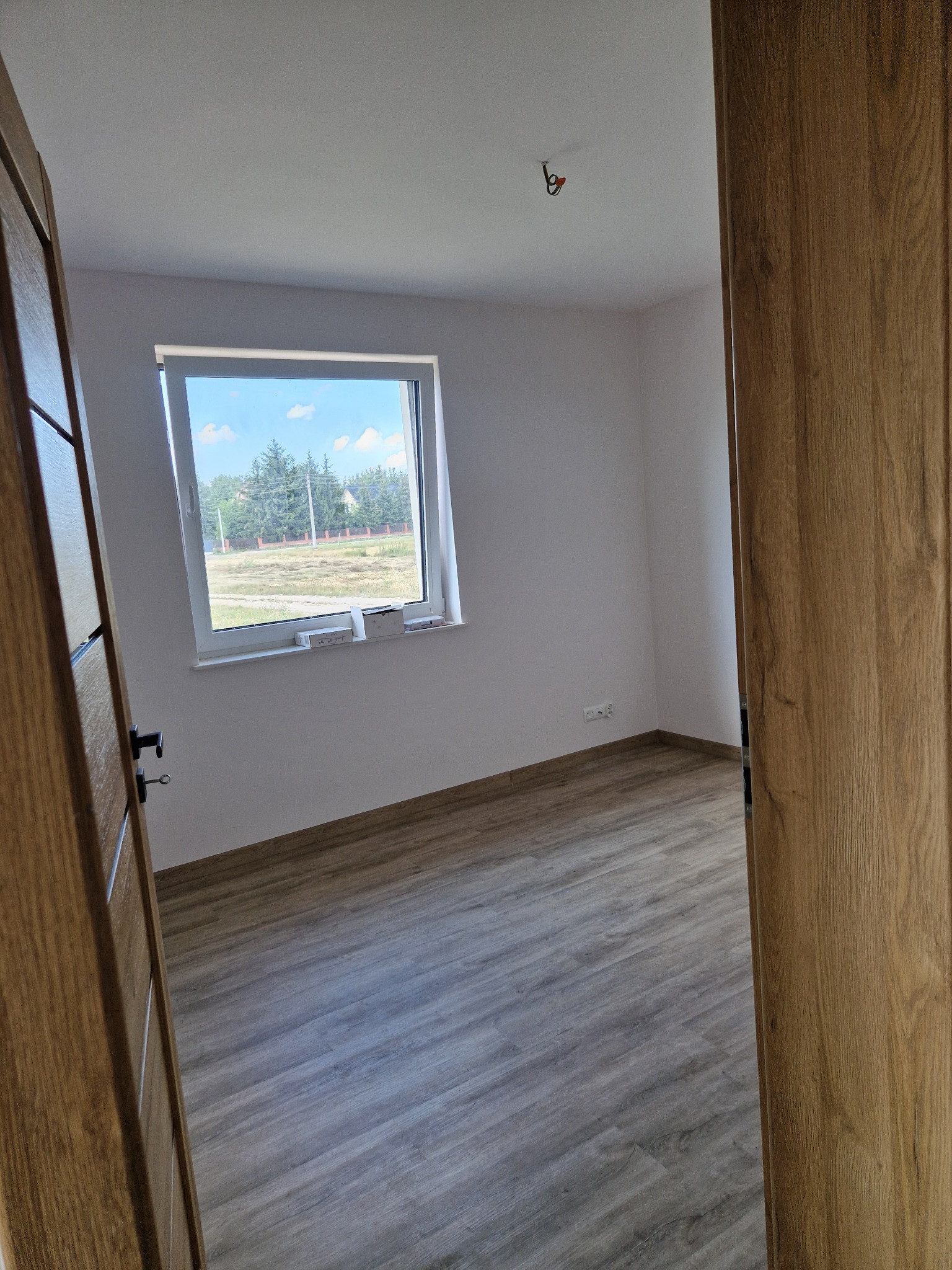 Pusty, jasny pokój z drewnianą podłogą i świeżo pomalowanymi ścianami, widok przez otwarte, drewniane drzwi, okno z widokiem na zewnątrz, przygotowany do dalszych prac wykończeniowych.