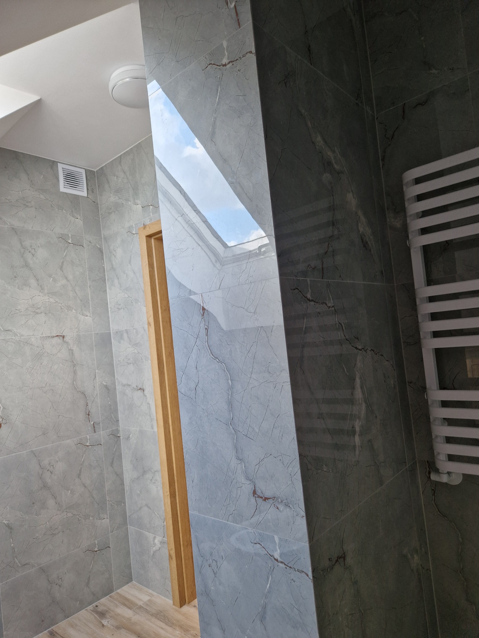 Wykończona łazienka z szarymi płytkami imitującymi marmur, drewnianą ościeżnicą i grzejnikiem drabinkowym, widoczny fragment okna dachowego.