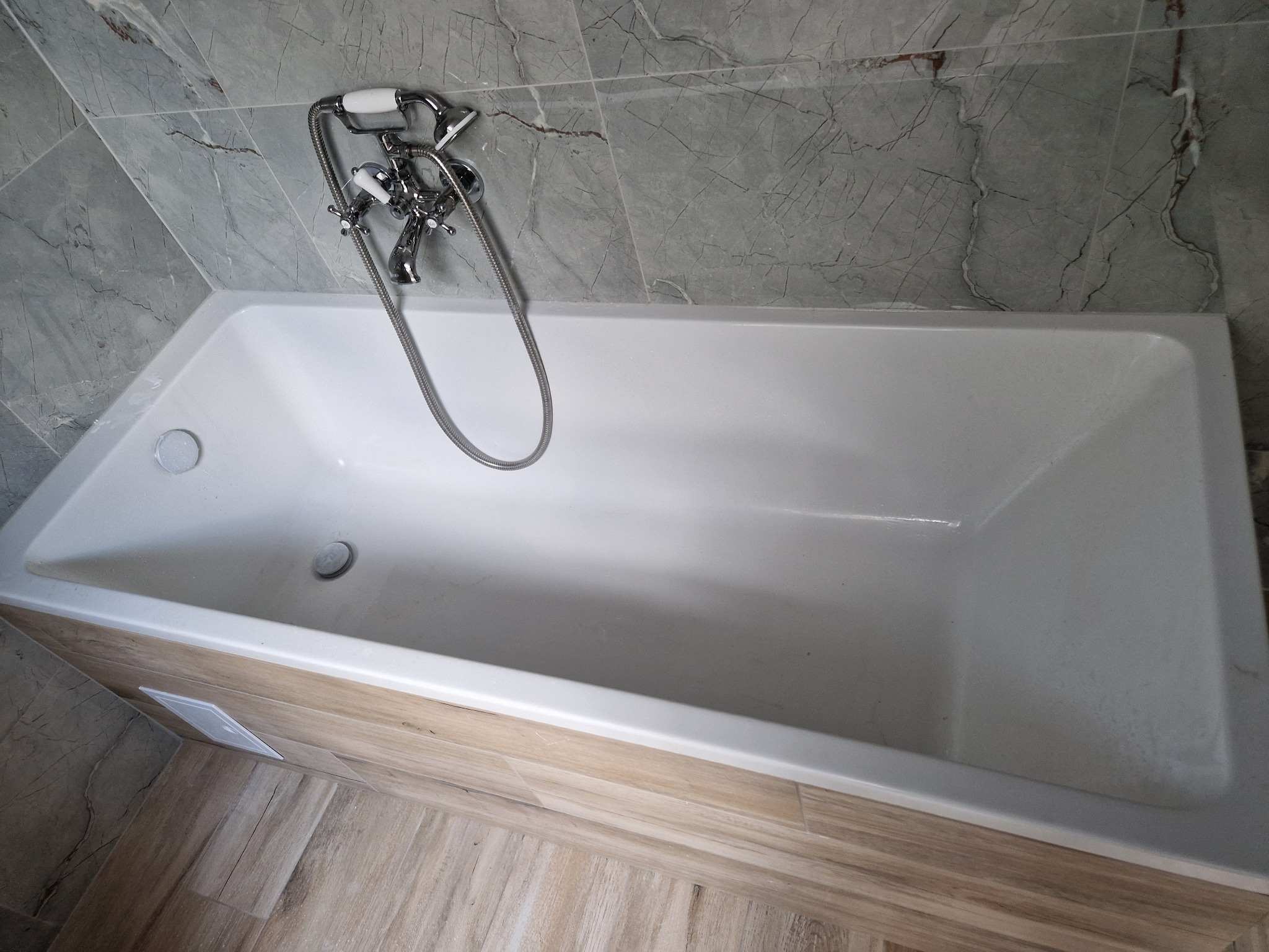 Wanna obudowana panelami imitującymi drewno, z chromowaną baterią i szarymi płytkami imitującymi marmur na ścianie, widok z góry.