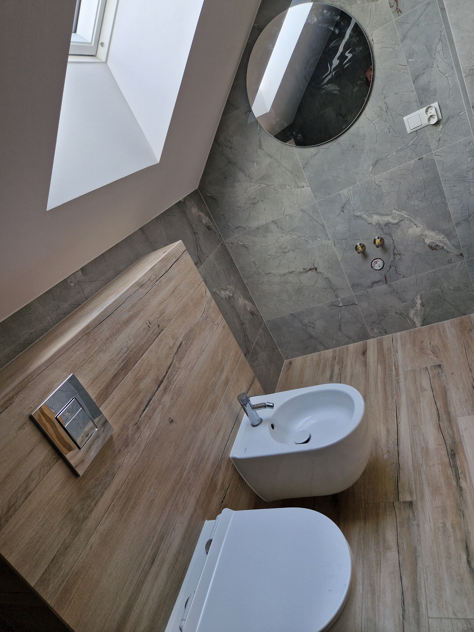 Nowoczesna łazienka na poddaszu z drewnopodobnymi płytkami, szarą marmurową ścianą, okrągłym lustrem i białą armaturą.