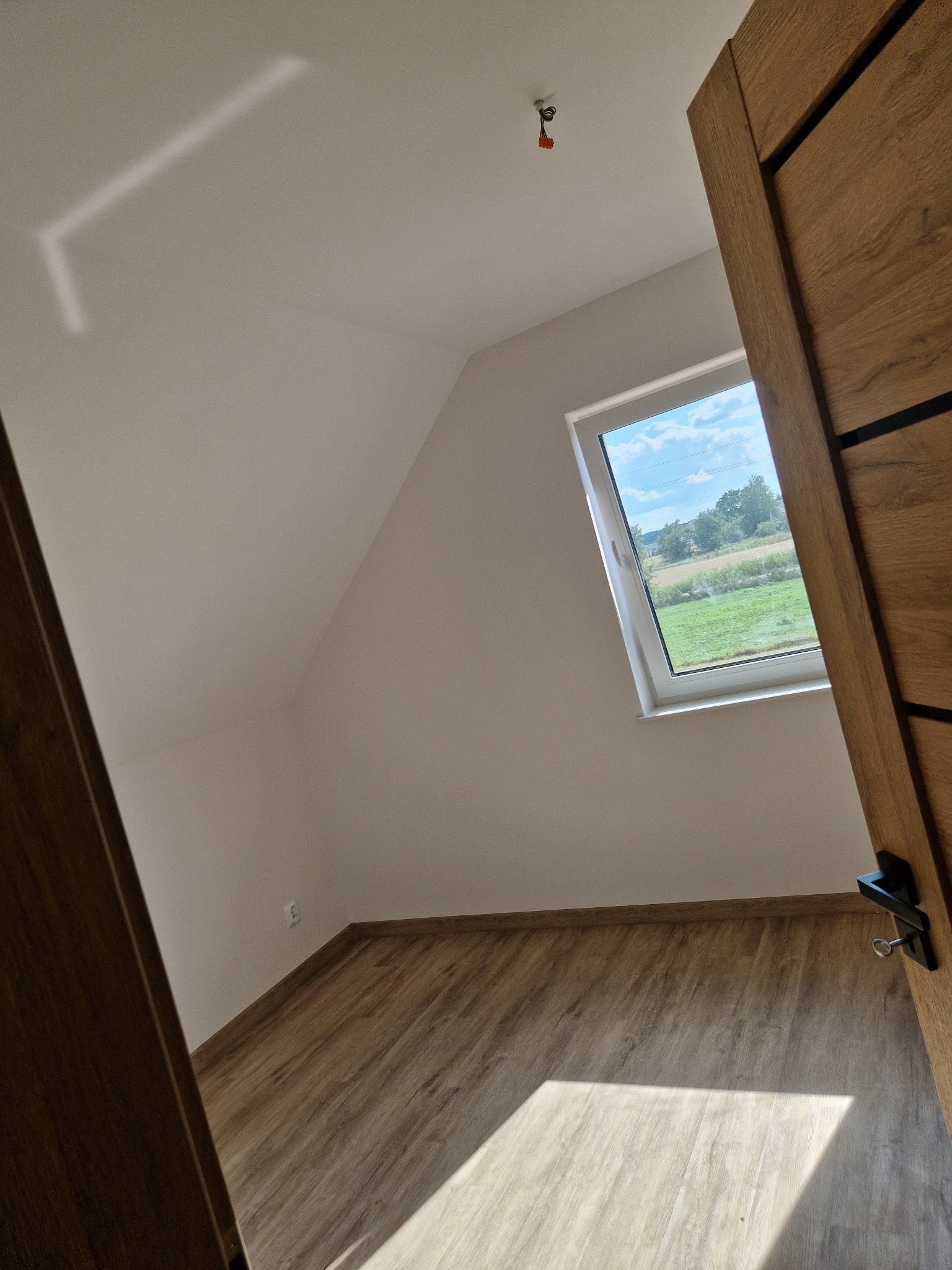 Poddasze z oknem dachowym i drewnopodobną podłogą, widok przez otwarte drzwi z drewnianą okleiną i czarną klamką, widoczne przewody oświetleniowe.