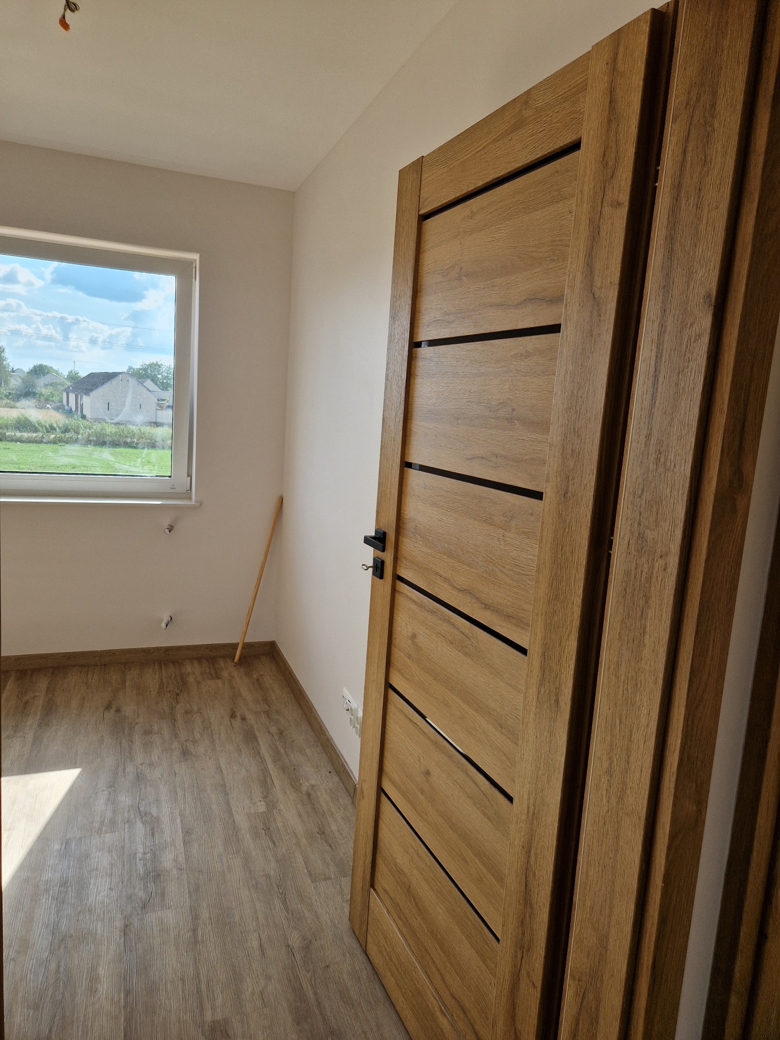 Nowe, drewniane drzwi wewnętrzne z czarnymi wstawkami, zamontowane w pokoju z panelami podłogowymi i widokiem na zewnątrz przez okno. Na ścianie widoczne wystające rury.